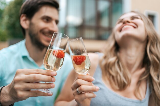 Glückliches Paarporträt, das zwei Gläser mit Sekt und Erdbeeren innen mit unscharfem Haus auf Hintergrund klirrt. Liebe feiern