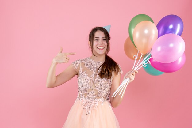 glückliches Partygirl mit Partykappe, das Luftballons hält, die mit Finger selbst auf Rosa zeigen