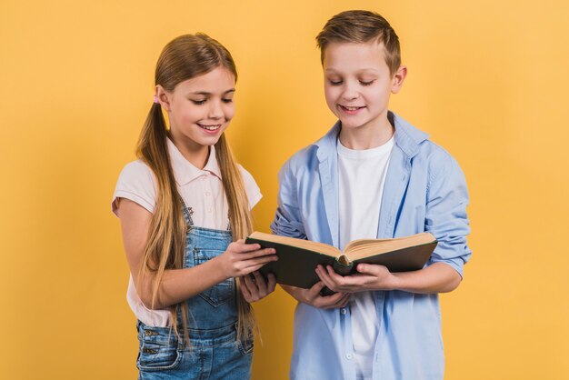 Glückliches Porträt des Jungen und des Mädchens, die das Buch stehen gegen gelben Hintergrund lesen