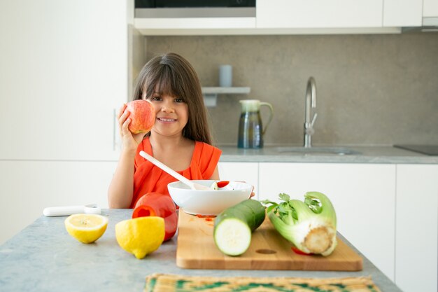 Glückliches positives Mädchen, das am Küchentisch mit geschnittenem frischem Gemüse steht und Apfel hält und zeigt, lächelnd, Kamera betrachtend. Gesundes Ernährungskonzept