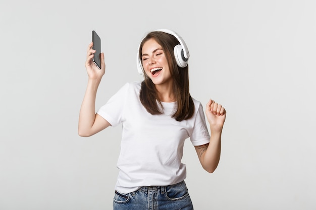 Glückliches schönes brünettes Mädchen tanzt und hört Musik in kabellosen Kopfhörern, hält Smartphone.