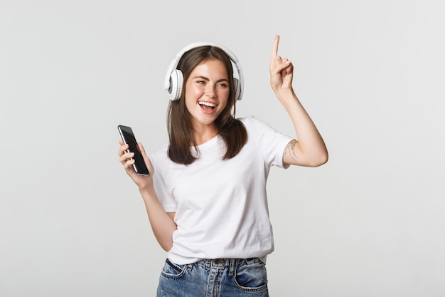 Glückliches schönes brünettes Mädchen tanzt und hört Musik in kabellosen Kopfhörern, hält Smartphone.