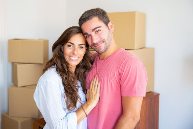 Glückliches schönes hispanisches Paar, das zwischen Kartonschachteln in ihrer neuen Wohnung steht, umarmt und Kamera betrachtet
