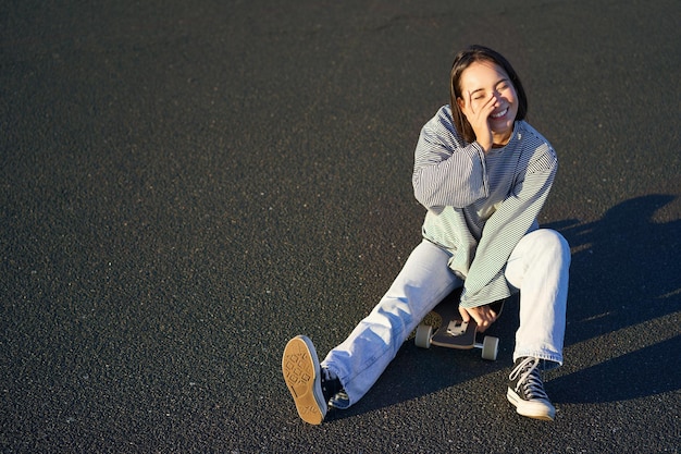 Glückliches schönes koreanisches jugendlich Mädchen sitzt auf ihrem Skateboard, das auf Longboard kreuzt und lässige Kleidung trägt