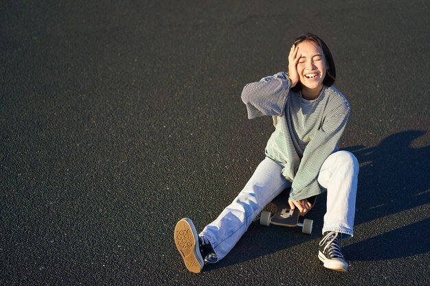 Glückliches schönes koreanisches jugendlich Mädchen sitzt auf ihrem Skateboard, das auf Longboard kreuzt und lässige Kleidung trägt