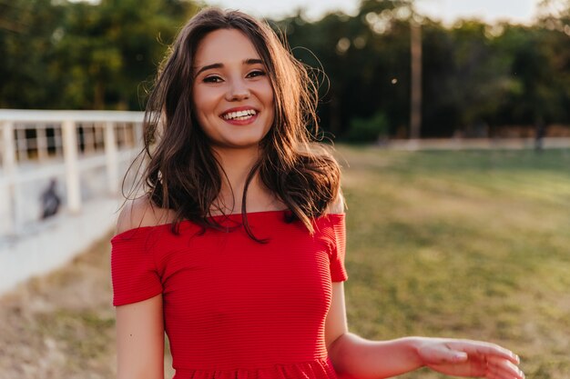 Glückliches schönes Mädchen, das auf Unschärfennatur mit Lächeln steht. Verträumte brünette Frau im roten Kleid, das mit aufrichtigem Lachen aufwirft