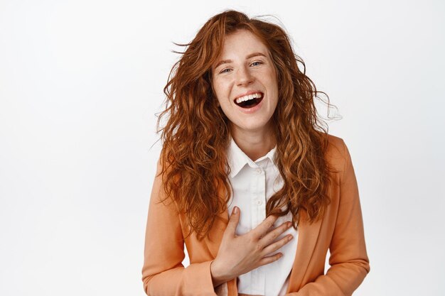 Glückliches schönes Mädchen mit lockigem rotem Haar, das lacht und lächelt und ehrliche Gefühle zeigt, die im Anzug vor weißem Hintergrund stehen