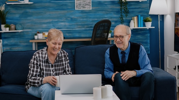 Glückliches Seniorenpaar winkt bei Videoanruf mit Neffen mit Laptop auf der Couch im Wohnzimmer