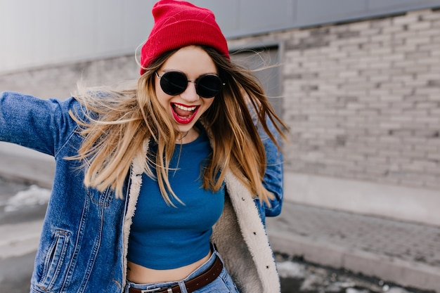 Glückliches siegreiches Mädchen in Jeans mit braunem Gürtel, das Spaß auf der Straße hat. Außenporträt der spektakulären kaukasischen Dame in der schwarzen Sonnenbrille, die auf Stadtwand tanzt.