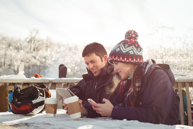 Glückliches Skifahrerpaar, das am Tisch sitzt, während Handy und digitales Tablet verwendet werden