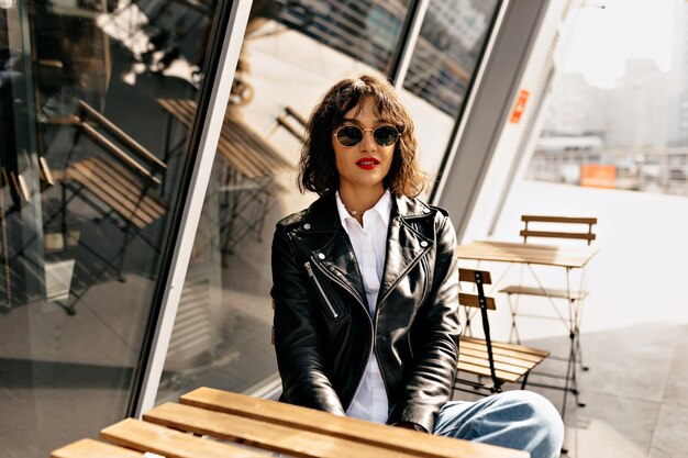 Glückliches stilvolles Mädchen mit kurzen Haaren, die Brille und Lederjacke mit roten Lippen tragen, die auf Sommerterrasse im Sonnenlicht ruhen