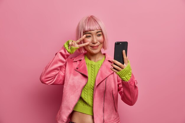 Glückliches stilvolles tausendjähriges Mädchen zeigt zwei Finger über Auge, Friedenszeichen, nimmt selfie, genießt ihre neue Frisur, gefärbtes Haar in Rosa