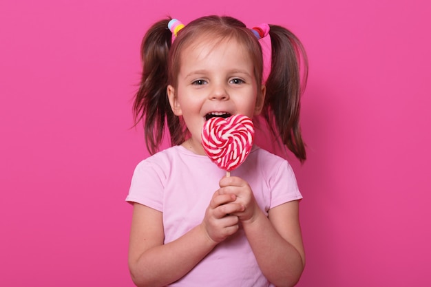 Glückliches süßes Mädchen trägt Rose T-Shirt, steht isoliert auf Rosa, hält hellen Lutscher in Händen. Fröhliches Kind mit geöffnetem Mund, das köstliche Süßigkeiten schmeckt. Konzept für Kindheit und Geschmack.