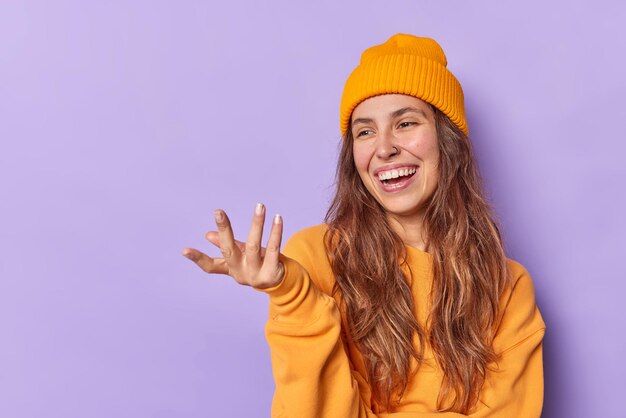 Glückliches Teenager-Mädchen hebt die Hand freudig auf etwas Lustiges trägt Hut und Pullover posiert vor violettem Hintergrund mit Leerzeichen für Ihre Werbeinhalte. Positives Konzept der menschlichen Emotionen