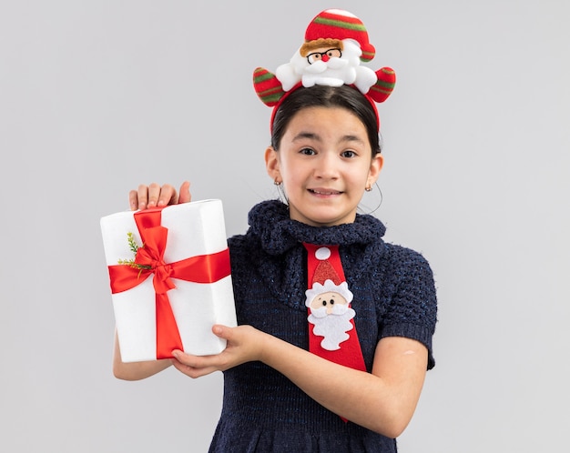 Glückliches und fröhliches kleines mädchen im strickkleid, das rote krawatte mit lustigem weihnachtsrand auf kopf hält, der weihnachtsgeschenk hält, das mit lächeln auf gesicht schaut