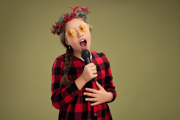 Glückliches und positives kleines Mädchen, das Weihnachtskranz im karierten Kleid hält, das Mikrofon singend steht über grünem Hintergrund hält