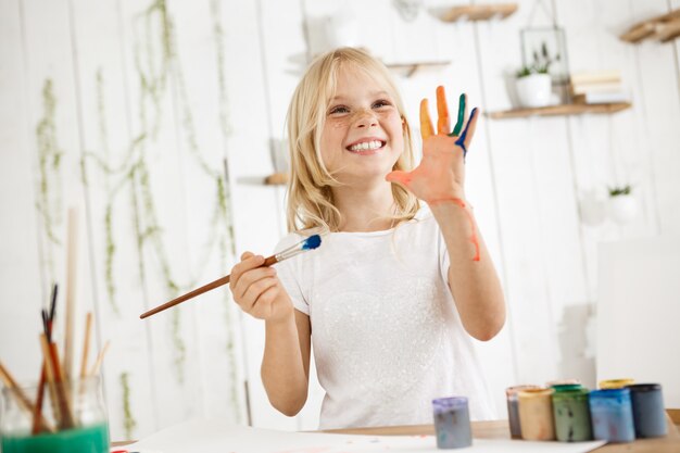 Glückliches und verspieltes süßes sommersprossiges blondes Mädchen, das in Weiß gekleidet ist, Pinsel in einer Hand hält und eine andere Hand zeigt, die sie mit Farbe durcheinander brachte.