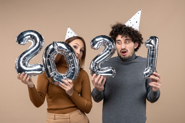 Glückliches verwirrtes junges Paar tragen Neujahrshut posiert für Kamera Mädchen zeigt und und Kerl mit und auf Grau