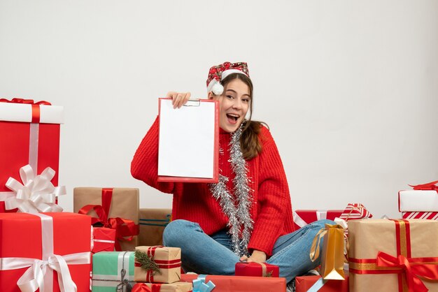 glückliches Weihnachtsmädchen mit Weihnachtsmütze, die Dokument zeigt, das um Geschenke auf Weiß sitzt