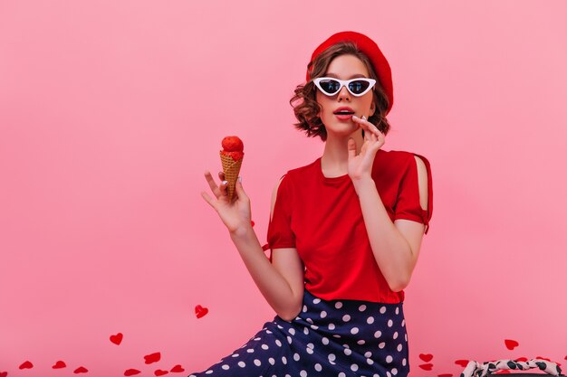 Glückliches weißes Mädchen in der eleganten Sonnenbrille, die Eiscreme isst. Schönes französisches weibliches Modell, das kaltes Dessert genießt.