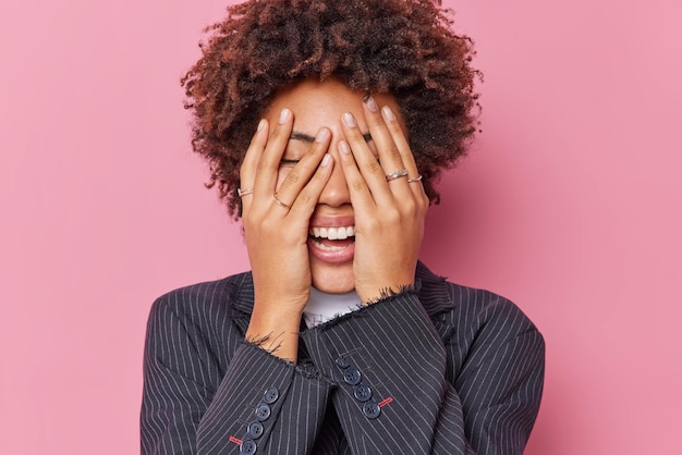 Kostenloses Foto glücks- und emotionskonzept fröhliche junge lockige frau versteckt gesicht mit händen kichert positiv versucht, ihre gefühle zu verbergen, die in einer formellen schwarzen jacke gekleidet sind, die über rosafarbenem studiohintergrund isoliert ist