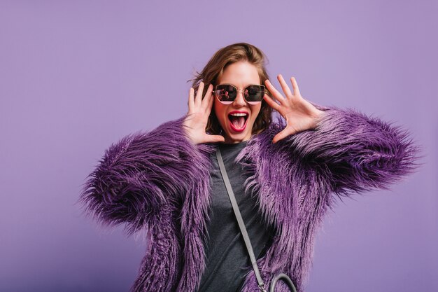 Glückselige Frau in grauer Kleidung und lila Jacke, die Spaß am Innen-Fotoshooting hat