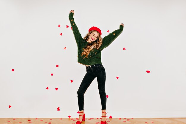 Glückselige Frau in roten Schuhen mit hohen Absätzen, die auf weißer Wand unter Herzkonfetti tanzen