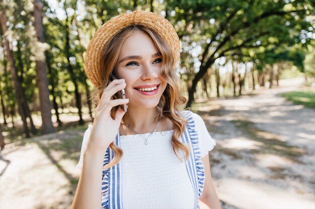 Glückselige junge Dame im trendigen Strohhut, der während des Telefongesprächs lächelt. Foto im Freien des erstaunlichen weißen Mädchens, das Freund anruft.