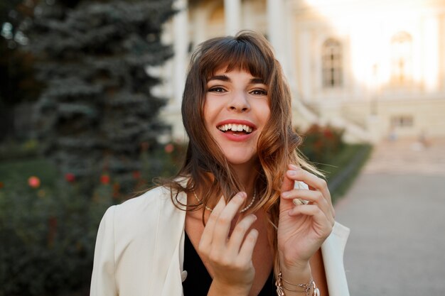 Glückselige lachende Frau, die mit Haaren spielt und uotumn Tag in der europäischen Stadt genießt. Eleganter Look.