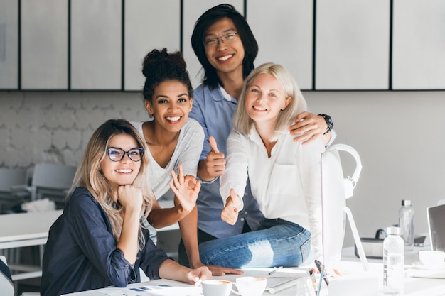 Kostenloses Foto glückseliger asiatischer programmierer, der weibliche blonde kollegin für foto und lächeln umarmt. innenporträt der fröhlichen afrikanischen büroangestellten, die neben europäischer freundin an ihrem arbeitsplatz aufwirft.