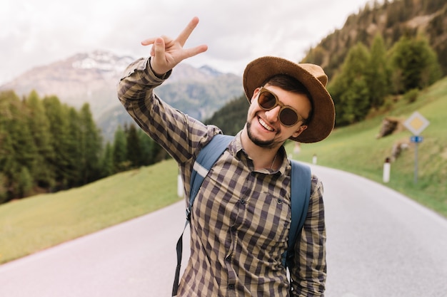 Glückseliger junger Mann in der trendigen Sonnenbrille, die mit Lächeln während der Wanderreise zu den italienischen Alpen aufwirft, die Friedenszeichen zeigen