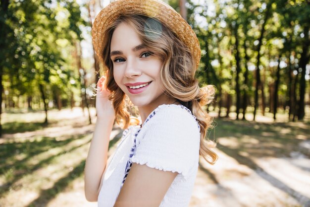 Glückseliges blondes Mädchen im Strohhut, das spielerisch im Wald aufwirft. Außenaufnahme der herrlichen Dame, die über Schulter auf Natur schaut.