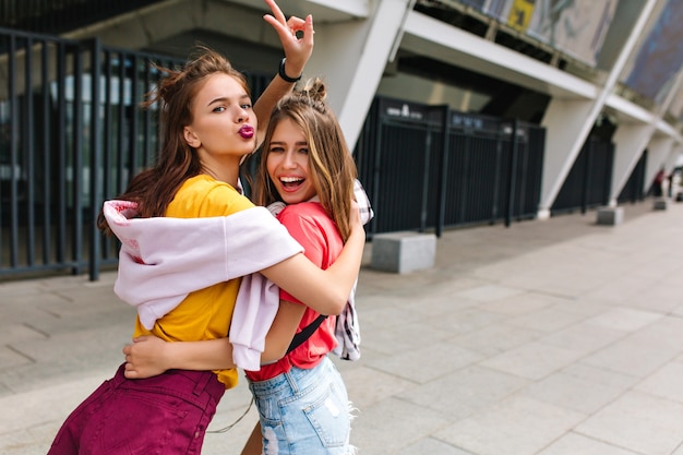 Glückseliges braunhaariges Mädchen in den trendigen lila Shorts, die mit Friedenszeichen aufwerfen und Gesichtsausdruck küssenden Freund umarmen