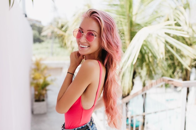 Glückseliges europäisches Mädchen mit schönem Lächeln, das an sonnigen Tagen auf Naturhintergrund lacht Fröhliche gebräunte Frau mit rosa Haaren, die Glück im exotischen Sommerresort ausdrückt