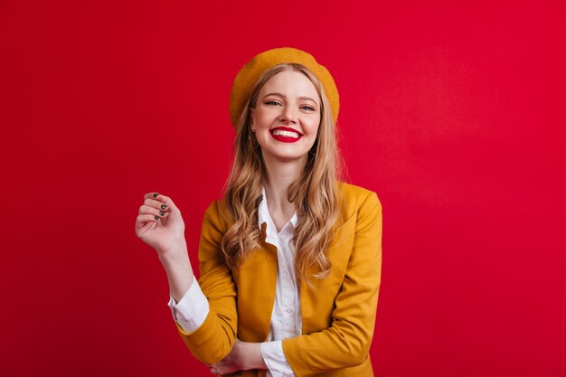 Glückseliges französisches weibliches Modell, das lacht. Vorderansicht des blonden Mädchens in der Baskenmütze lokalisiert auf roter Wand.