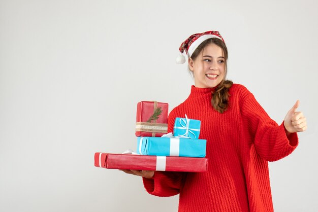 Glückseliges Mädchen der Vorderansicht mit der Weihnachtsmütze, die Daumen hoch Zeichen macht, das ihr Weihnachtsgeschenk hält