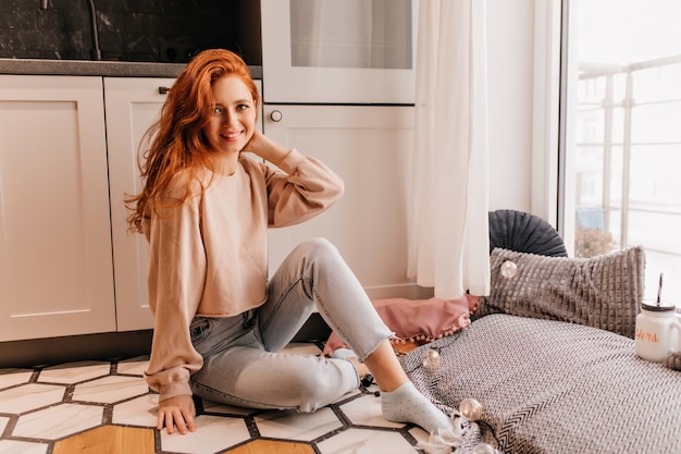 Glückseliges Mädchen des Ingwers, das spielerisch zu Hause posiert. Innenfoto der gewinnenden jungen Dame in den Jeans.