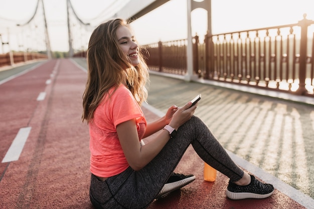 Glückseliges Mädchen in den schwarzen Turnschuhen, die an der Schlackenbahn sitzen und lachen. Emotionale junge Frau, die mit Smartphone und Flasche Saft aufwirft.