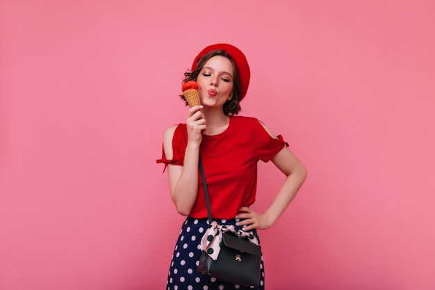 Glückseliges Mädchen mit der schwarzen Handtasche, die Eiscreme genießt. Ekstatisches weibliches Modell in der Baskenmütze, die mit Nachtisch aufwirft.