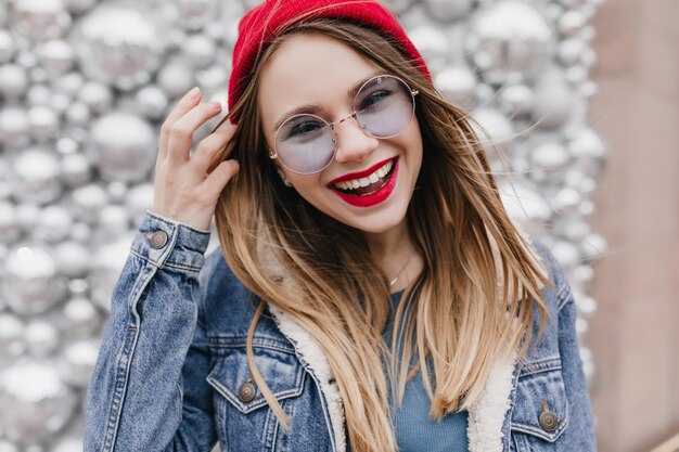 Glückseliges weißes Mädchen mit schönem Lächeln, das auf funkelndem Hintergrund posiert und mit ihren Haaren spielt Porträt eines glücklich lachenden weiblichen Models in trendiger Jeansjacke