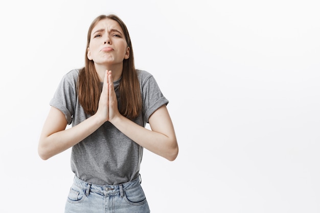 Gott, bitte. Attraktives junges kaukasisches Studentenmädchen mit mittleren Haarlängen im lässigen Outfit, das Palmen zusammenpresst, nach oben schaut und um Hilfe in einer schwierigen Situation beim Lernen bittet.