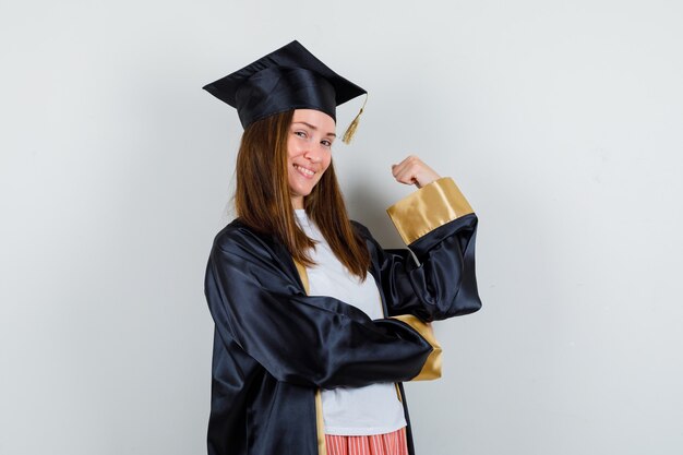 Graduierte Frau, die Siegergeste in der Freizeitkleidung, Uniform zeigt und lustig schaut, Vorderansicht.