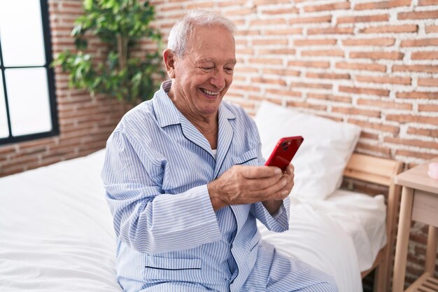 Grauhaariger Mann mittleren Alters mit Smartphone, der im Schlafzimmer auf dem Bett sitzt