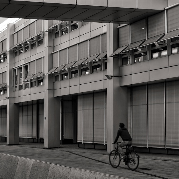Graustufenaufnahme eines Mannes, der auf dem Bürgersteig neben einem Gebäude mit vielen Fenstern Fahrrad fährt