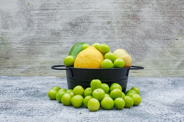 Greengages, Birnen und Zitrone in einem schwarzen Topf auf grungy blauem und grauem Holz. Seitenansicht.