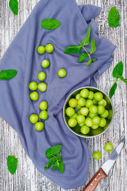 Greengages mit Blättern in einem Metalltopf mit Messer flach auf grauem Holz und Picknicktuch liegen