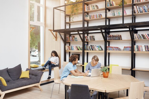 Große moderne Bibliothek am Morgen. Zwei Leute sitzen und schauen in Laptop-Monitor, der über Startprojekt spricht. Mädchen, das auf dem Fensterbank-Lesebuch auf dem Smartphone sitzt, verbringt Zeit vor dem Studium.