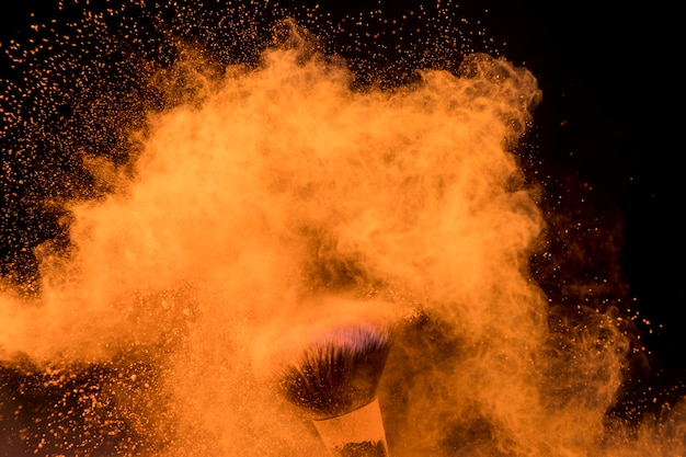 Große Wolke des orange Pulvers um Make-upbürste auf dunklem Hintergrund