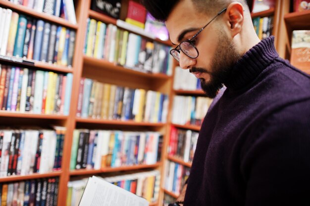 Großer, intelligenter arabischer Student trägt einen violetten Rollkragenpullover und eine Brille in der Bibliothek und liest ein Buch