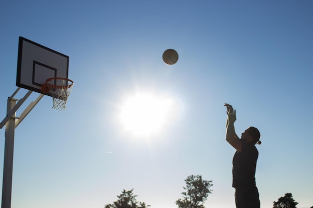 Großer Mann, der an einem sonnigen Tag im Freien auf dem Sportplatz Basketball spielt. Mann wirft Basketball in Korb vor Zenit-Himmelshintergrund. Aktiver Lebensstil, Sport, Motivationskonzept